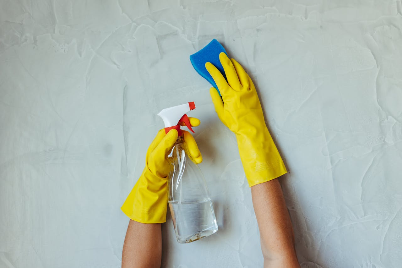 hero-img-02 Close-up of hands in yellow gloves cleaning a wall with a spray bottle and sponge.
